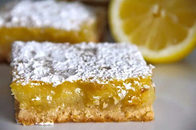 A close up of a lemon bar with the rest of the dessert in the background.